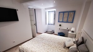 A bedroom with a large bed, a TV mounted on the wall, a window, a small plant, and a decorative wall panel.