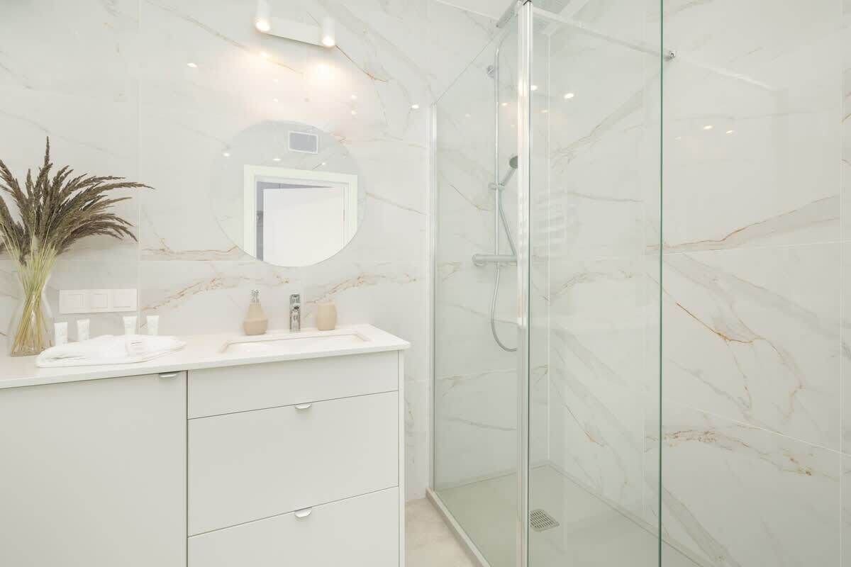 Un baño moderno con pared de mármol, cabina de ducha de vidrio y tocador blanco con espejo y artículos de aseo.