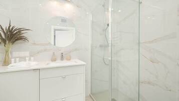 Un baño moderno con pared de mármol, cabina de ducha de vidrio y tocador blanco con espejo y artículos de aseo.