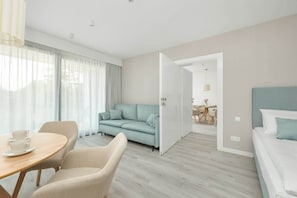 A modern hotel room with a sofa, a dining table with chairs, a bed, and a view through an open door.