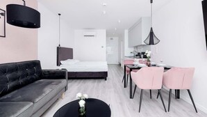 Ein modernes Hotelzimmer mit einem schwarzen Sofa, einem Esstisch mit rosa Stühlen, einem schwarzen Couchtisch und einem Bett mit weißer Bettwäsche.