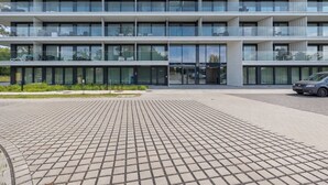 Ein mehrstöckiges Gebäude mit modernem Design, das Balkone mit Glasgeländern, große Fenster und einen gepflasterten Eingangsbereich aufweist.