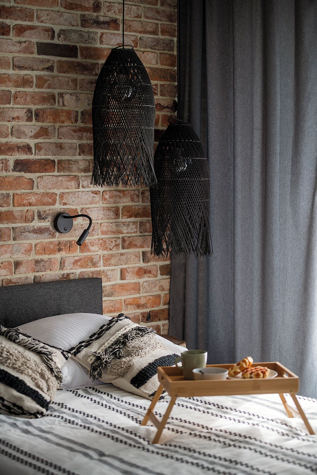A bedroom with a brick wall, a bed with patterned bedding, two hanging black lamps, and a tray with a cup and pastry.