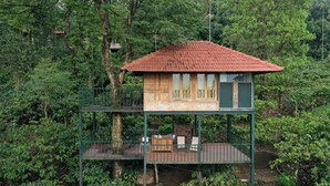Une maison en bois avec un toit rouge, surélevée sur des piliers, entourée d’une forêt dense.