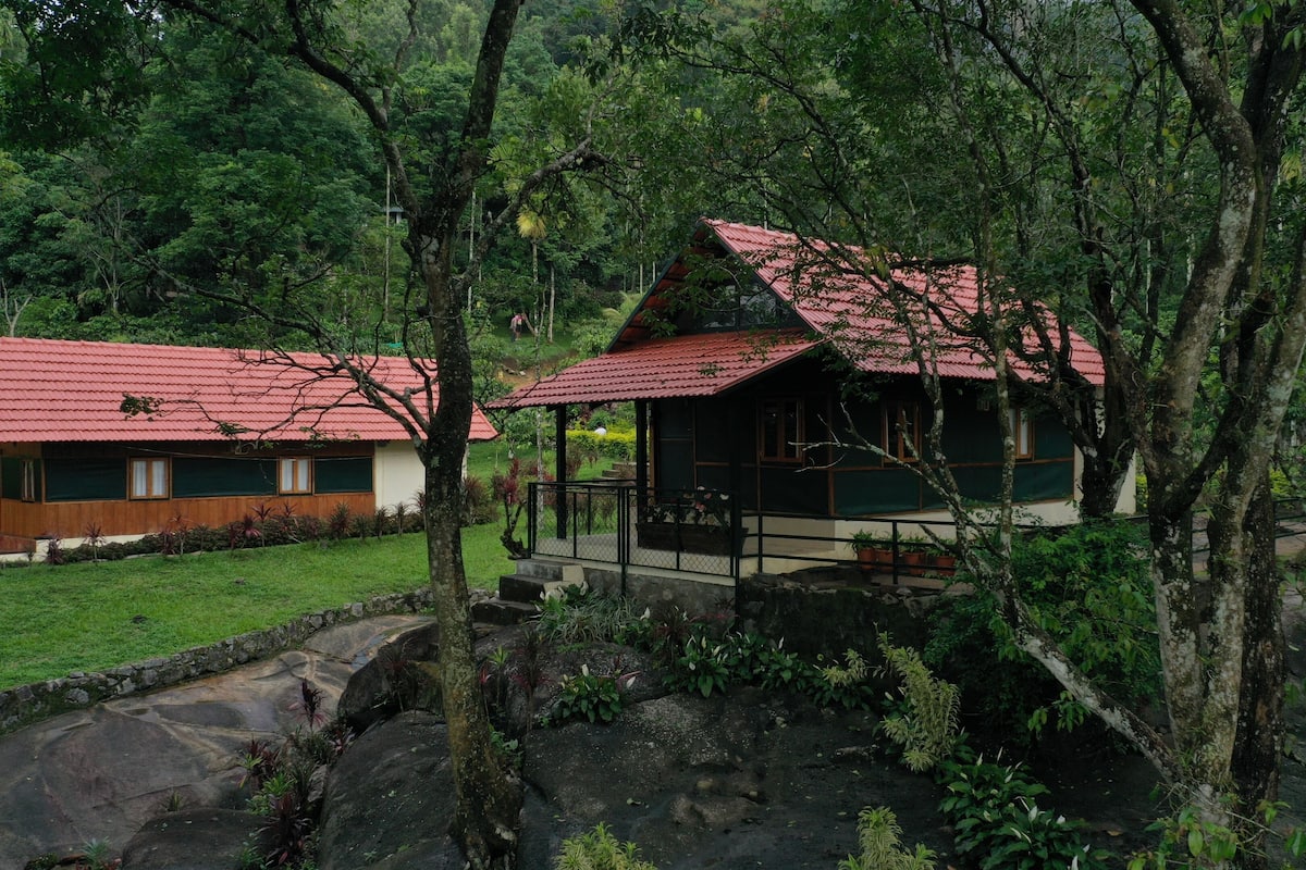 A rustic cabin with a red roof, surrounded by lush greenery and rocky terrain.