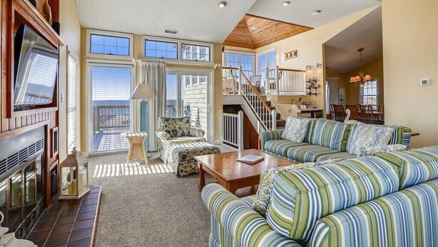 A spacious living room with a fireplace, striped sofas, a wooden ceiling fan, and a staircase leading to an upper level. - Waters, Ships Watch Community in Duck NC (Duck)