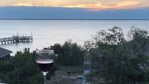 A glass of red wine on a balcony overlooking a calm body of water with a dock and a small building in the background. - Waters, Ships Watch Community in Duck NC (Duck)