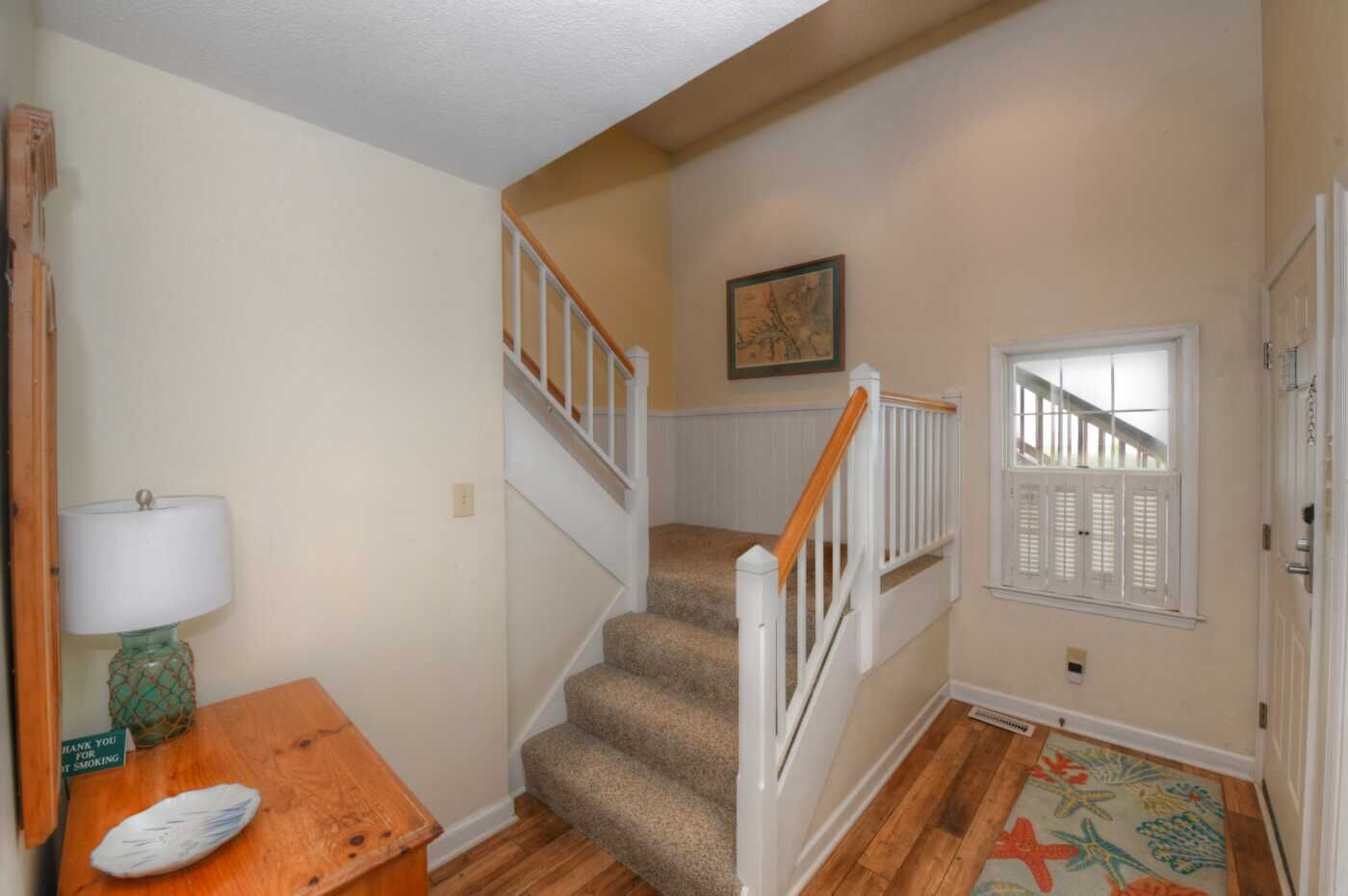 Un escalier avec une rampe en bois et des balustrades blanches, des marches recouvertes de moquette, et une table en bois sur laquelle se trouvent une lampe et un panneau.