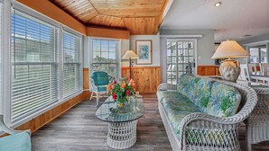 A spacious room with large windows, a wicker sofa, a round glass table, and a chair. - Waters, Ships Watch Community in Duck NC (Duck)
