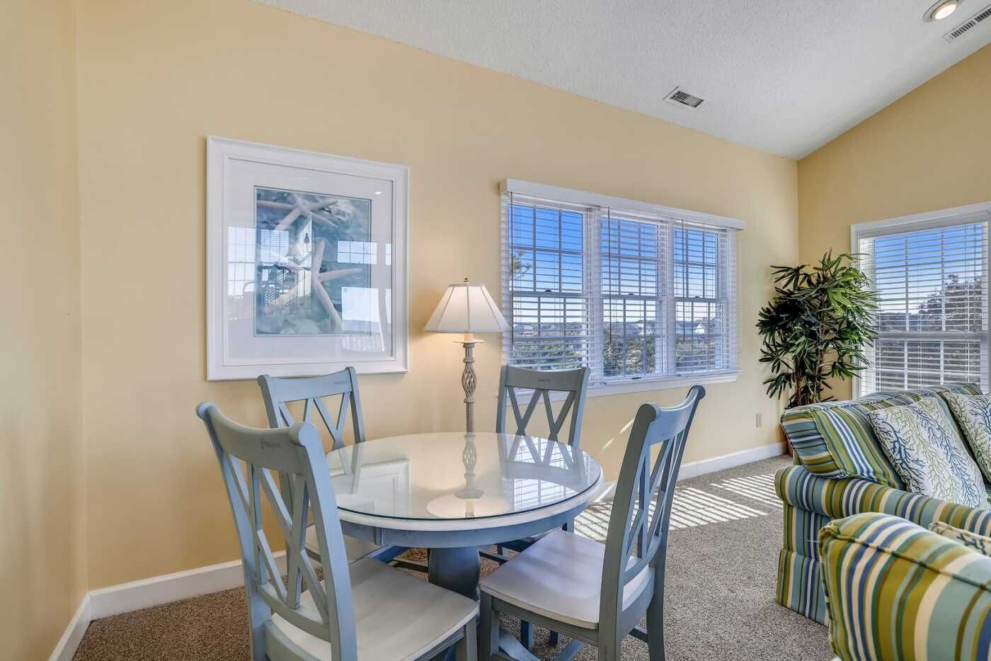 A dining area with a round table, four chairs, a lamp, and two windows with blinds.