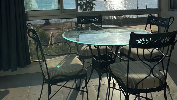Ein Zimmer mit Glastisch und Stühlen, großen Fenstern mit Blick auf Meer und Berge sowie einem Fliesenboden.