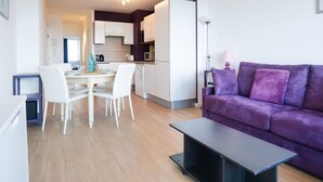 Una sala de estar moderna con un sofá morado, un comedor con sillas blancas y una cocina con armarios blancos.