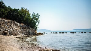 A rocky shoreline with a stone wall and trees, leading to calm water and distant mountains. - Red Eirth Kastel Stafilic (Kastela)