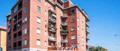 Un hotel de varios pisos con balcones, una entrada a nivel de calle y un cielo azul despejado.