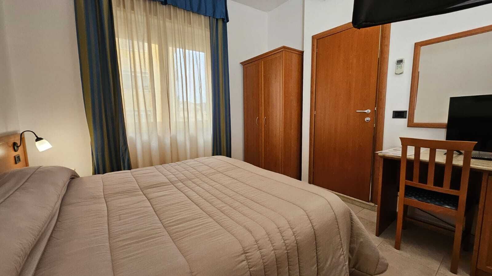 A hotel room with a bed, a desk, a chair, a wardrobe, and a television.