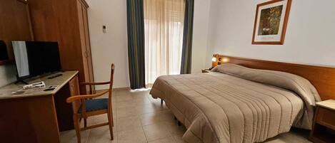 Una habitación de hotel con cama, escritorio con computadora, silla, ventana con cortinas y un cuadro en la pared.