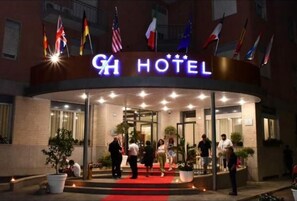 Exterior de un hotel con alfombra roja en la entrada, banderas y un letrero que dice 'GH HOTEL'.
