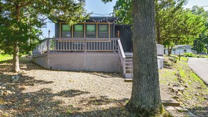 A small, two-story building with a balcony and a covered porch, surrounded by trees and gravel.