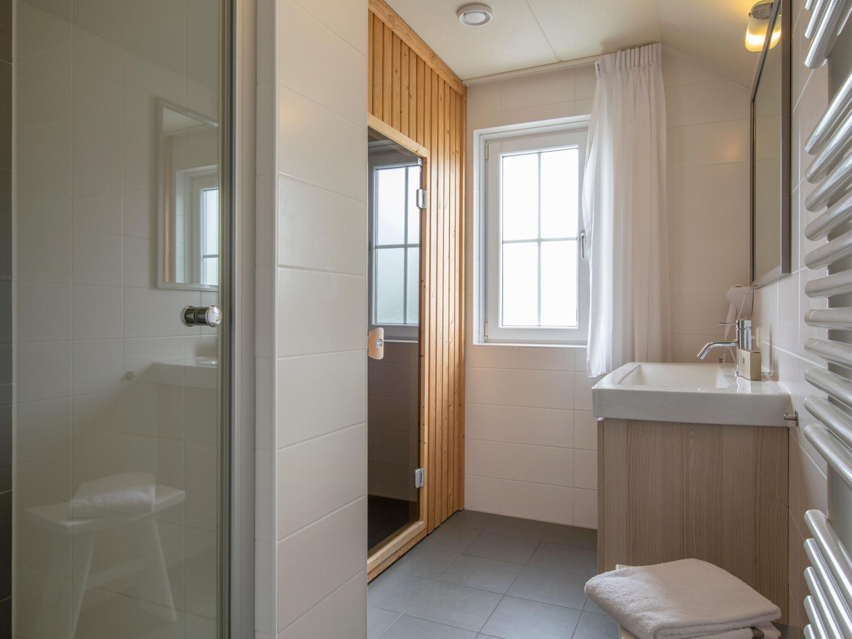 A modern bathroom with a glass shower enclosure, a wooden vanity, a white sink, and a window with curtains.