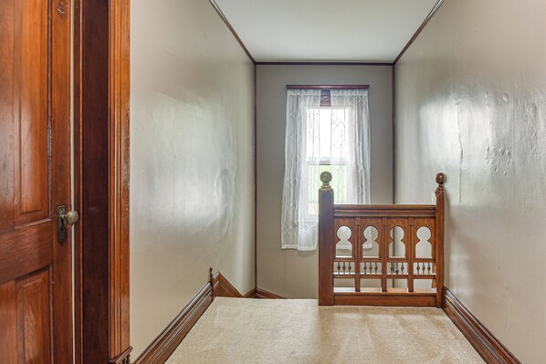 A narrow hallway with a wooden door on the left, a window with a sheer curtain, and a wooden bed frame at the end.