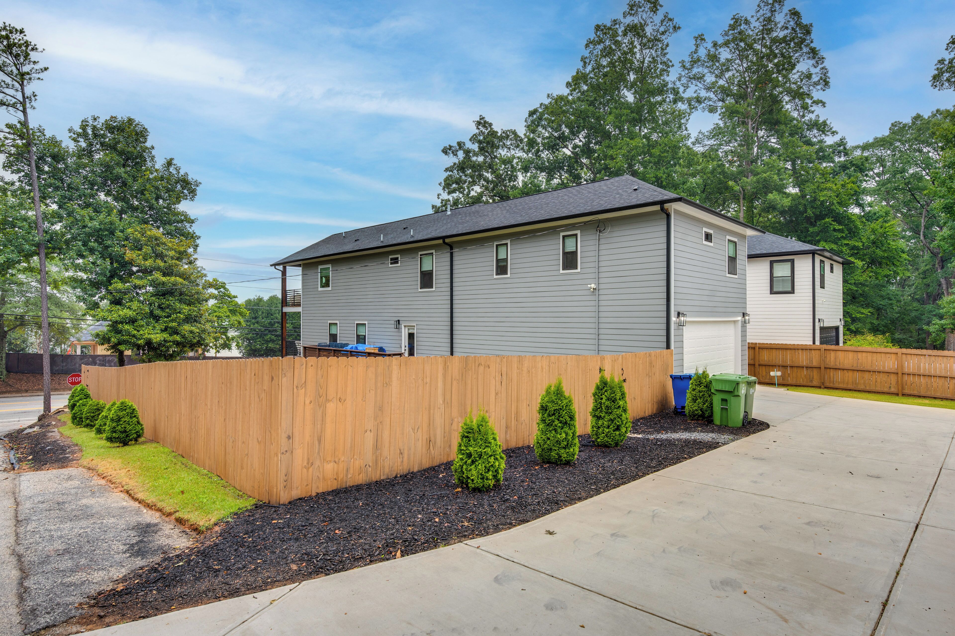 A two-story residential building with a grey siding exterior, a wooden fence, and a landscaped front yard with trimmed bushes.