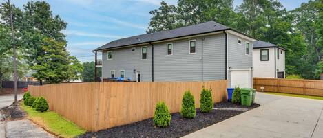 A two-story residential building with a grey siding exterior, a wooden fence, and a landscaped front yard with trimmed bushes.