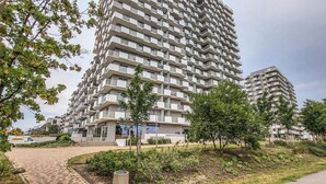 A modern high-rise building with a uniform facade, multiple balconies, and a paved entrance area with greenery.