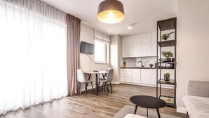 A modern living room with a dining area, wooden flooring, a sofa, a coffee table, and a kitchen with white cabinets and a plant shelf.