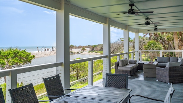 Ein überdachter Außenbereich mit Korbmöbeln, einem Tisch und Blick auf Strand und Meer.