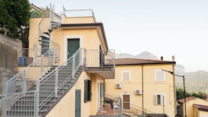 A yellow building with a spiral staircase leading to a balcony. There is a small outdoor seating area with a table and chairs.