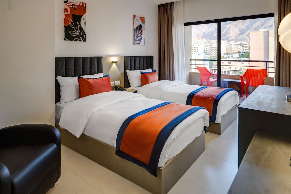 Ein Hotelzimmer mit zwei Betten, einem Schreibtisch und einem Balkon mit roten Stühlen.