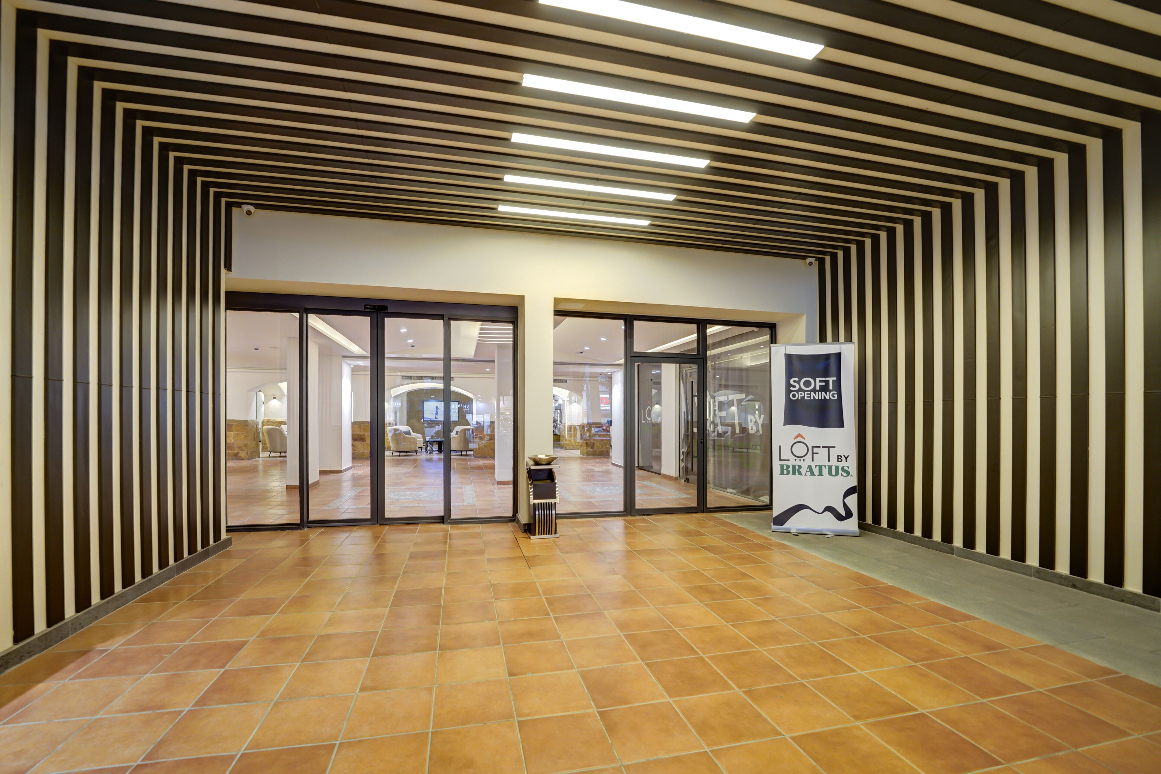 A modern hotel lobby with a geometric patterned ceiling, a tiled floor, and a glass door entrance.