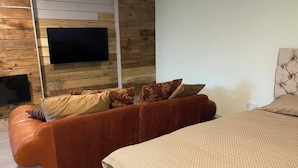 Une chambre moderne avec un lambris en bois, un téléviseur à écran plat, un canapé en cuir marron agrémenté de coussins à motifs et un lit recouvert d’une courtepointe texturée.