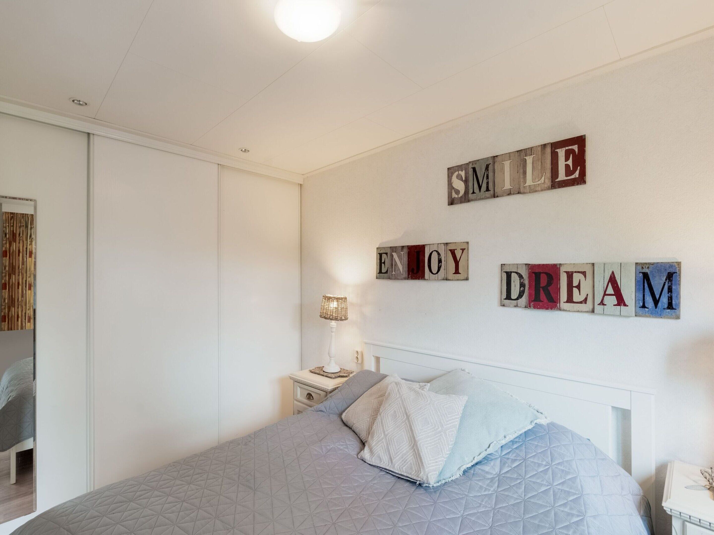 Een slaapkamer met een bed, kussens, een nachtkastje, een lamp en een motiverende muurdecoratie.