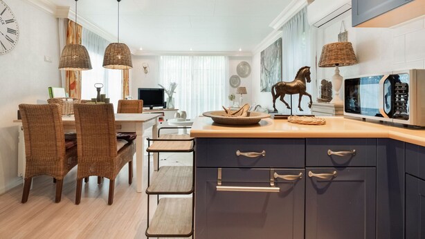 A modern kitchen with dark cabinets, a wooden dining table, wicker chairs, and a mounted TV. - Holiday Home in Garderen by Veluwe (Garderen)