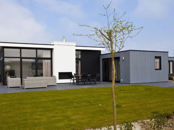 A modern house with a large glass window, a patio with outdoor furniture, and a small tree in the yard.