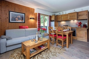 A cozy living room with a sofa, a coffee table with books, a dining area with chairs, and a kitchen with wooden cabinets.