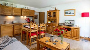 Uma sala de estar aconchegante com um sofá, uma mesa de jantar de madeira e cadeiras. Há uma área de cozinha com armários de madeira e uma televisão.