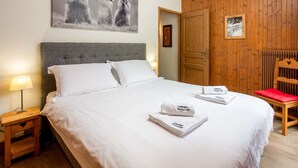 Cama bem arrumada com lençóis e toalhas brancos, cabeceira de madeira, mesa de cabeceira com abajur e uma cadeira de madeira com almofada vermelha.