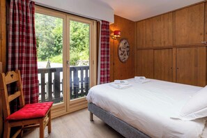 Un dormitorio con una cama, una silla, un armario de madera y vistas a través de un ventanal.