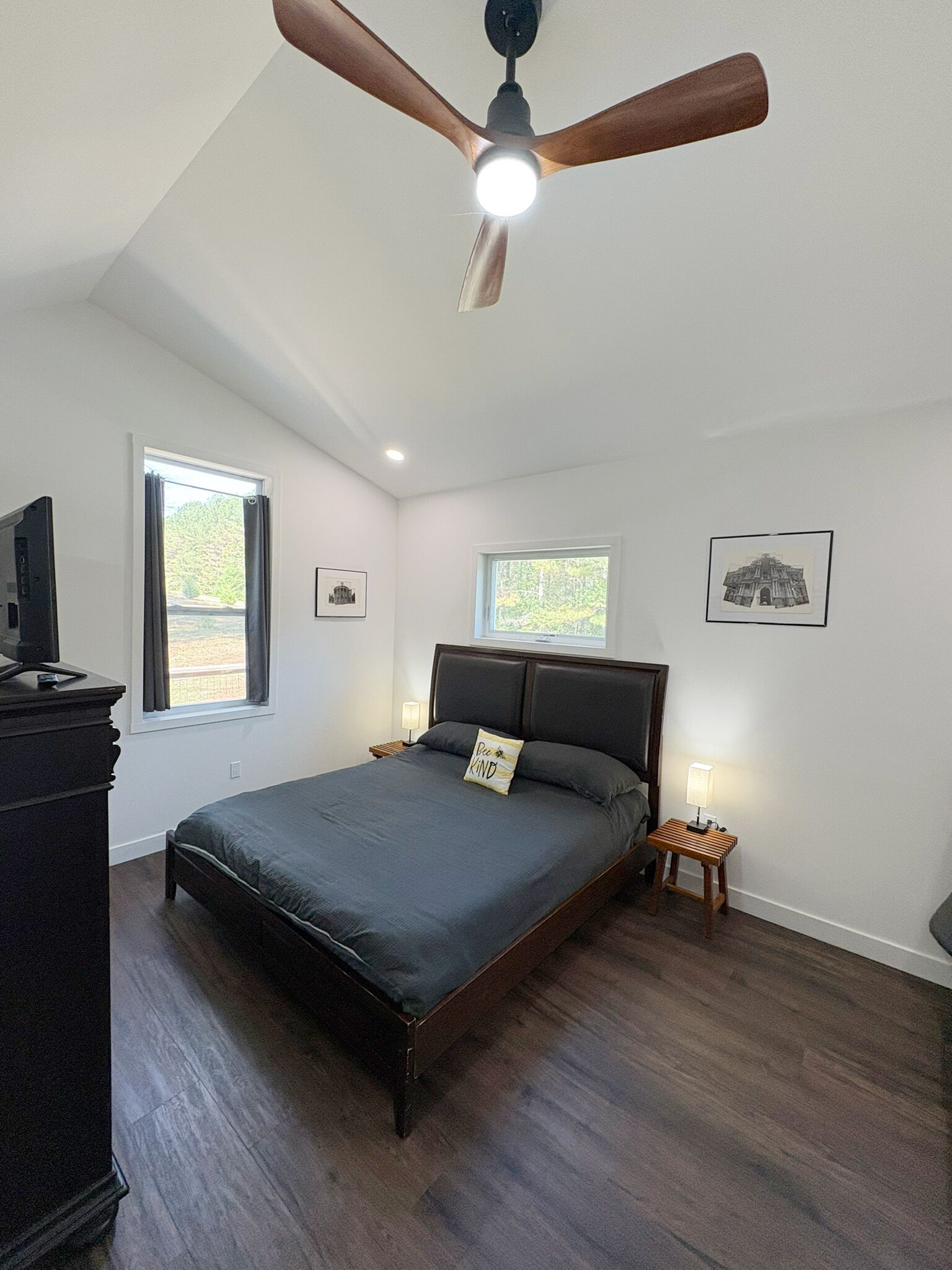 Une chambre à coucher comprenant un lit, un ventilateur de plafond, une table de chevet avec une lampe, une commode et deux fenêtres.