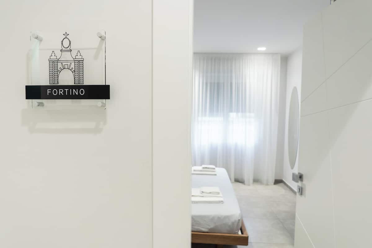 A hotel room with a bed, a window with sheer curtains, a wall-mounted sign with a fort-like drawing, and a mirror.