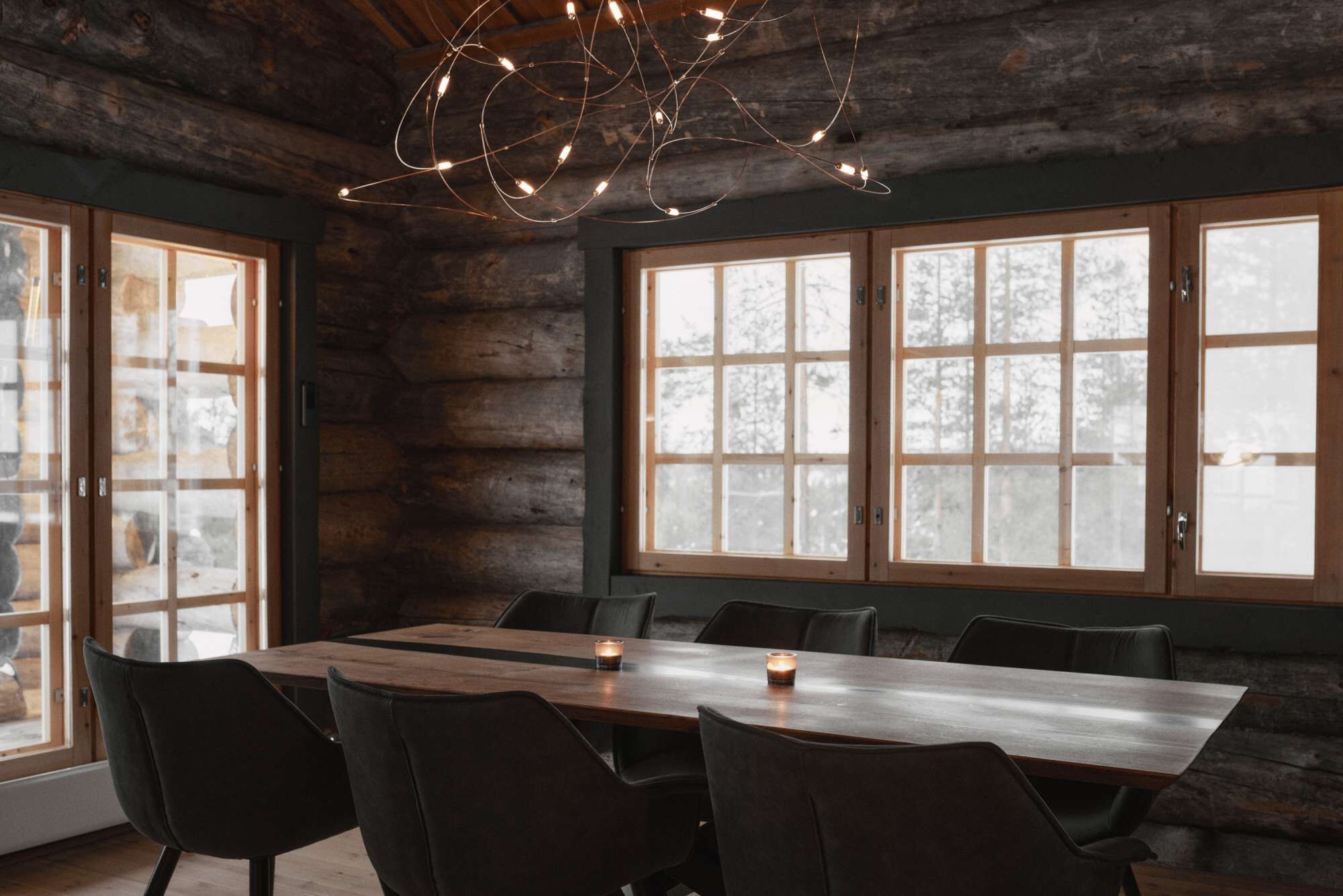 Un intérieur rustique en bois, avec une longue table à manger, de nombreuses chaises et de grandes fenêtres dotées de panneaux de verre dépoli.
