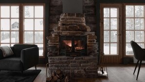 Una sala de estar acogedora con chimenea de piedra, dos ventanas amplias y un sofá de color oscuro.