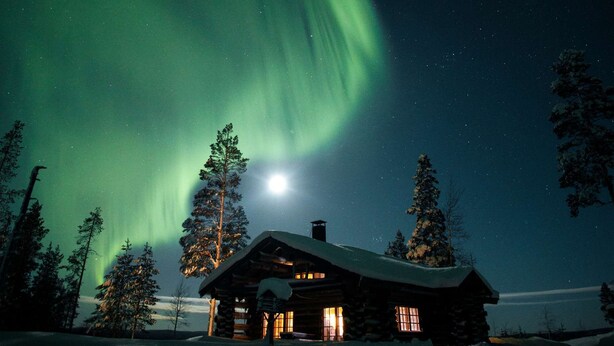 Una cabaña de troncos con techo nevado, rodeada de árboles, bajo un cielo nocturno con auroras boreales.