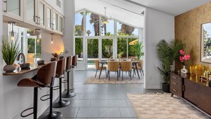 Une cuisine moderne avec des tabourets de bar, un coin repas avec une table et des chaises, de grandes fenêtres et une vue sur la verdure.