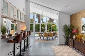 A modern kitchen with bar stools, a dining area with a table and chairs, large windows, and a view of greenery.