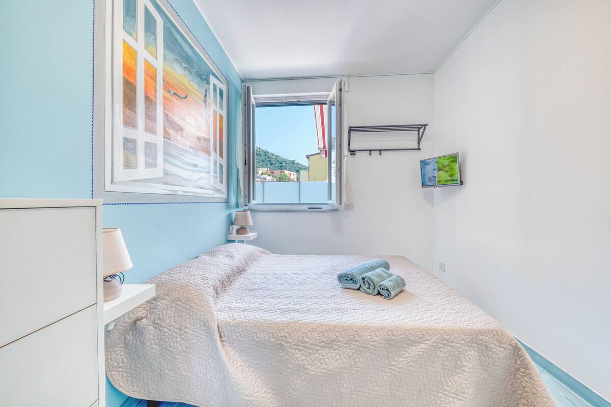A small, neatly arranged bedroom with a bed, a window, a lamp, and a painting on the wall.