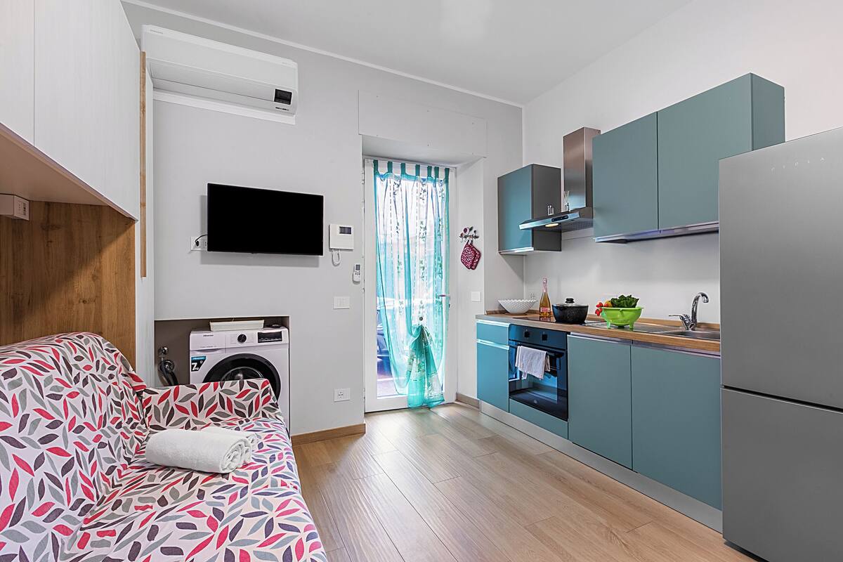 A compact living space with a sofa, a washing machine, a kitchen with teal cabinets, and a flat-screen TV mounted on the wall.