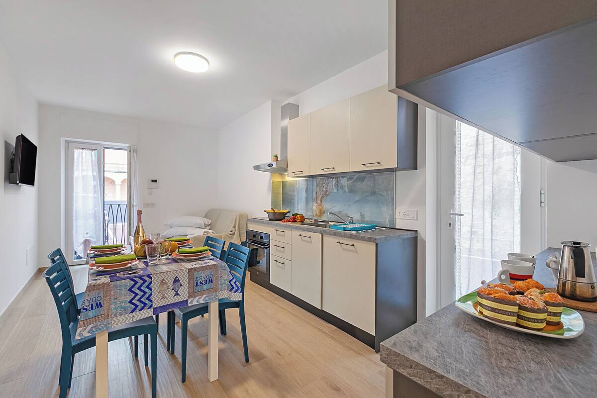 A modern kitchen with a dining area, featuring a table with chairs, a sink, and a countertop with a plate of pastries.
